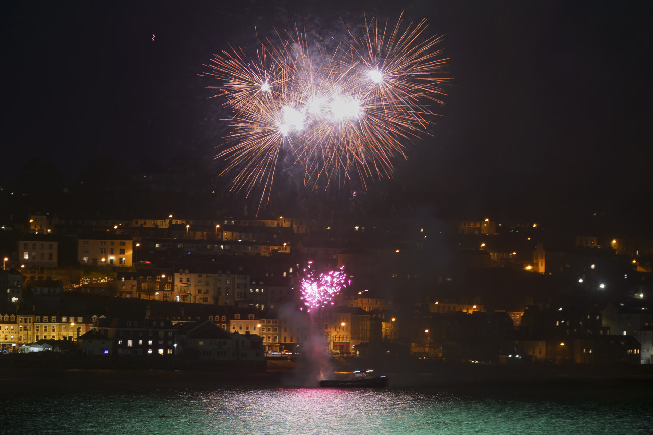 Fireworks light up the night sky over Cobh. The light reflects on the water.