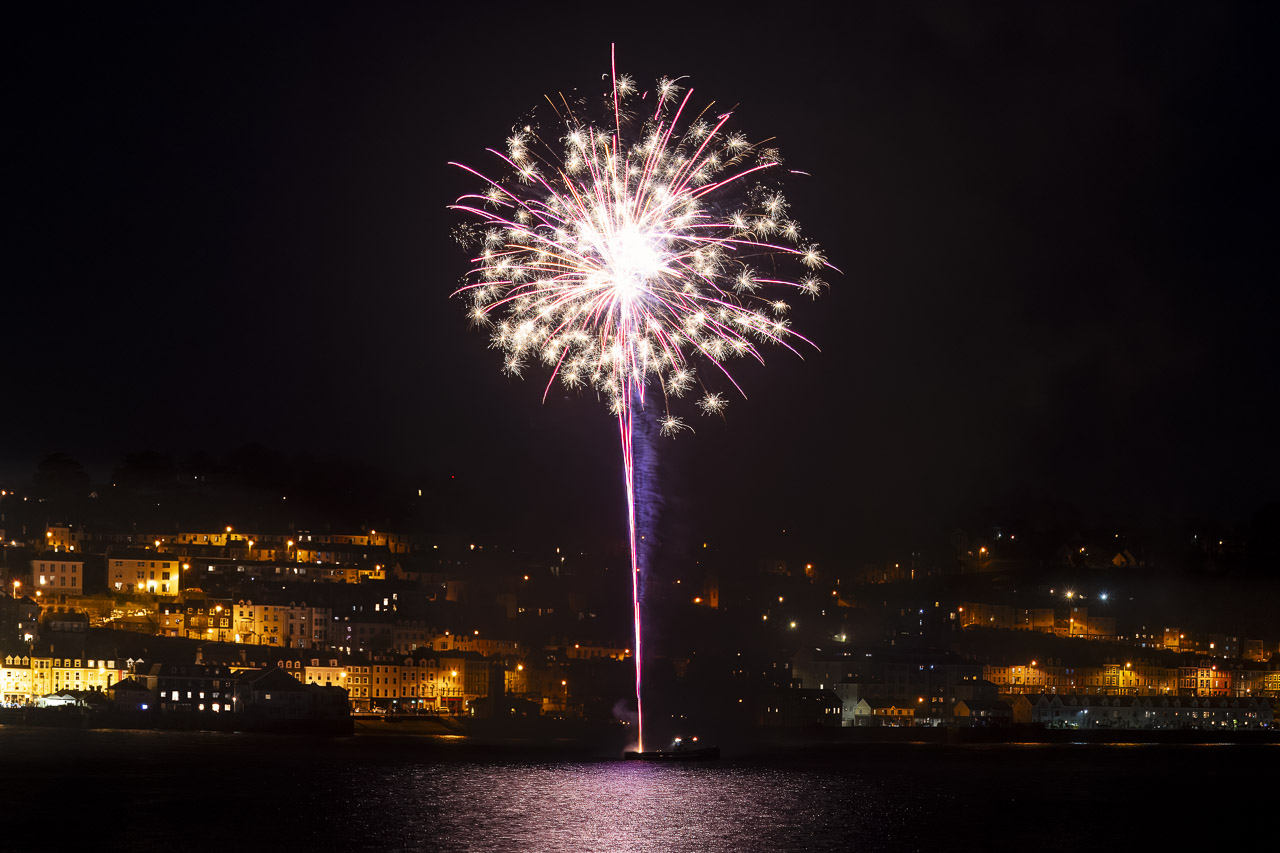 Fireworks light up the night sky over Cobh. The light reflects on the water.