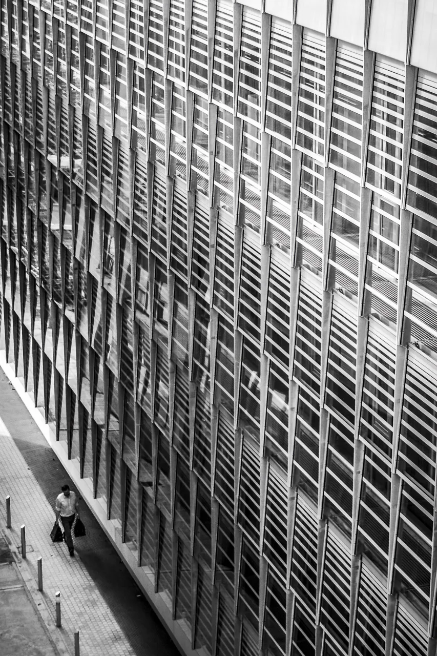 A man walking down an alley next to an office building with large windows.
He's carrying two bags, probably full of shopping.