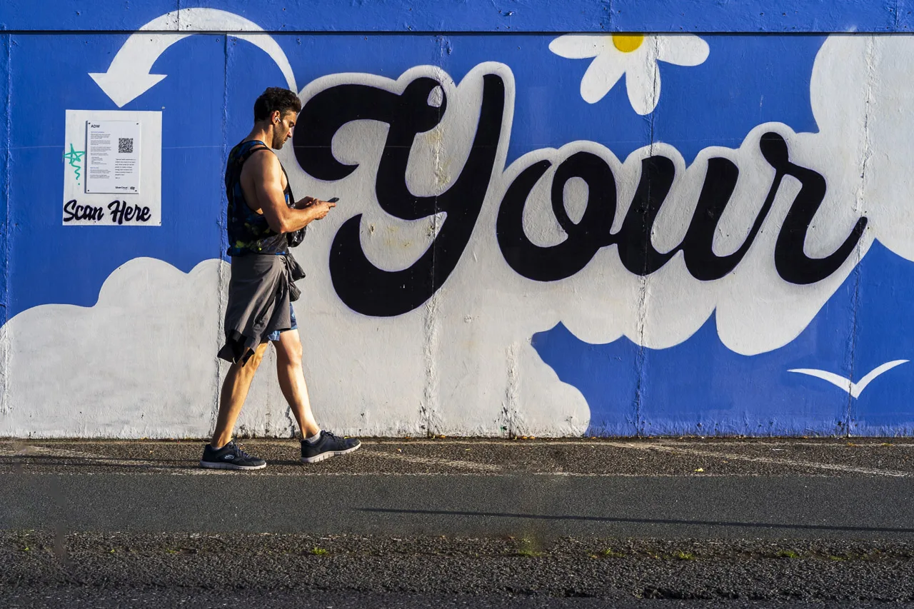 A man walking next to the hoarding around a building site with the word "Your" spray painted on it.