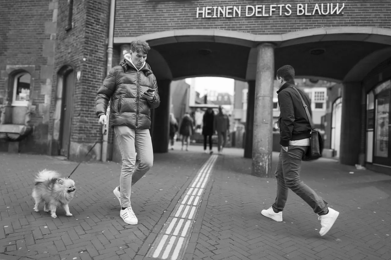 A man walking his dog is engrossed in his phone, while another man walks towards him.
In the background is a building with the words, "Heinen Delfts Blauw".
There is a tunnel through the building and people can be seen walking away and towards the viewer.