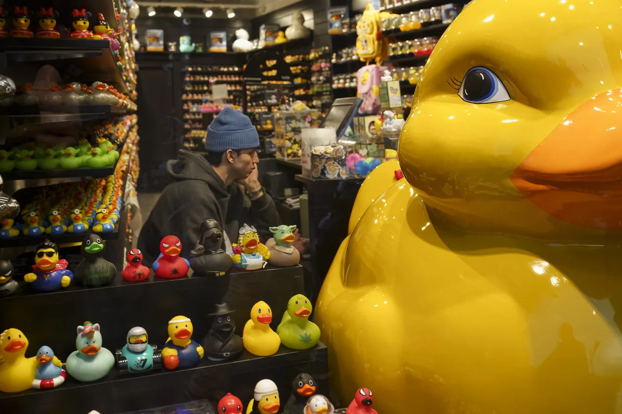 A bored looking man behind a counter in a shop full of rubber ducks, including a huge one to the right of the photo.