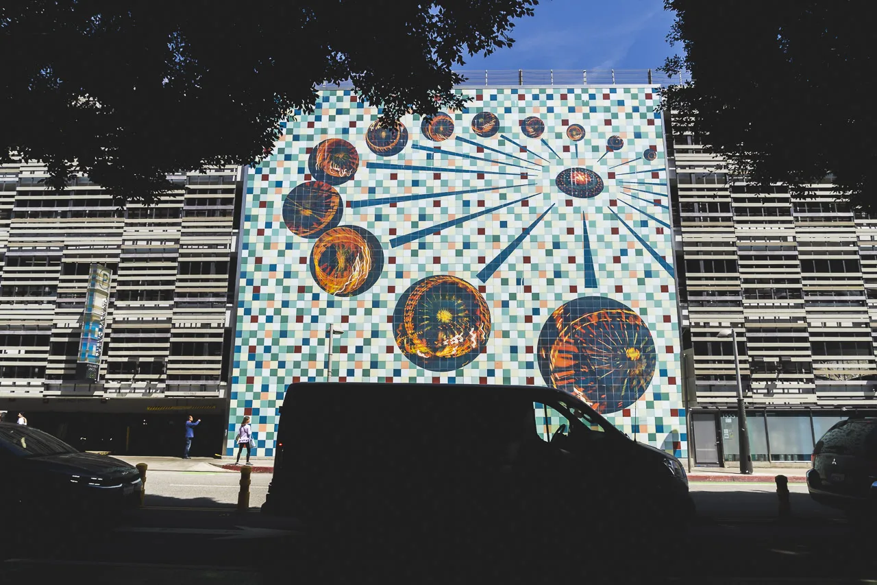 A mural called Wheels in Santa Monica, framed by trees and a parked van. The mural is in the shape of a wheel with circles at the end of each spoke.