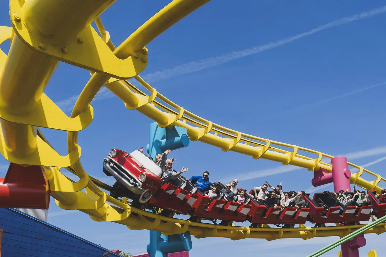 A section of a roller coaster with the car and people on it hurtling towards the camera. Some people have their arms raised. Others laughing, others grimly hanging on.