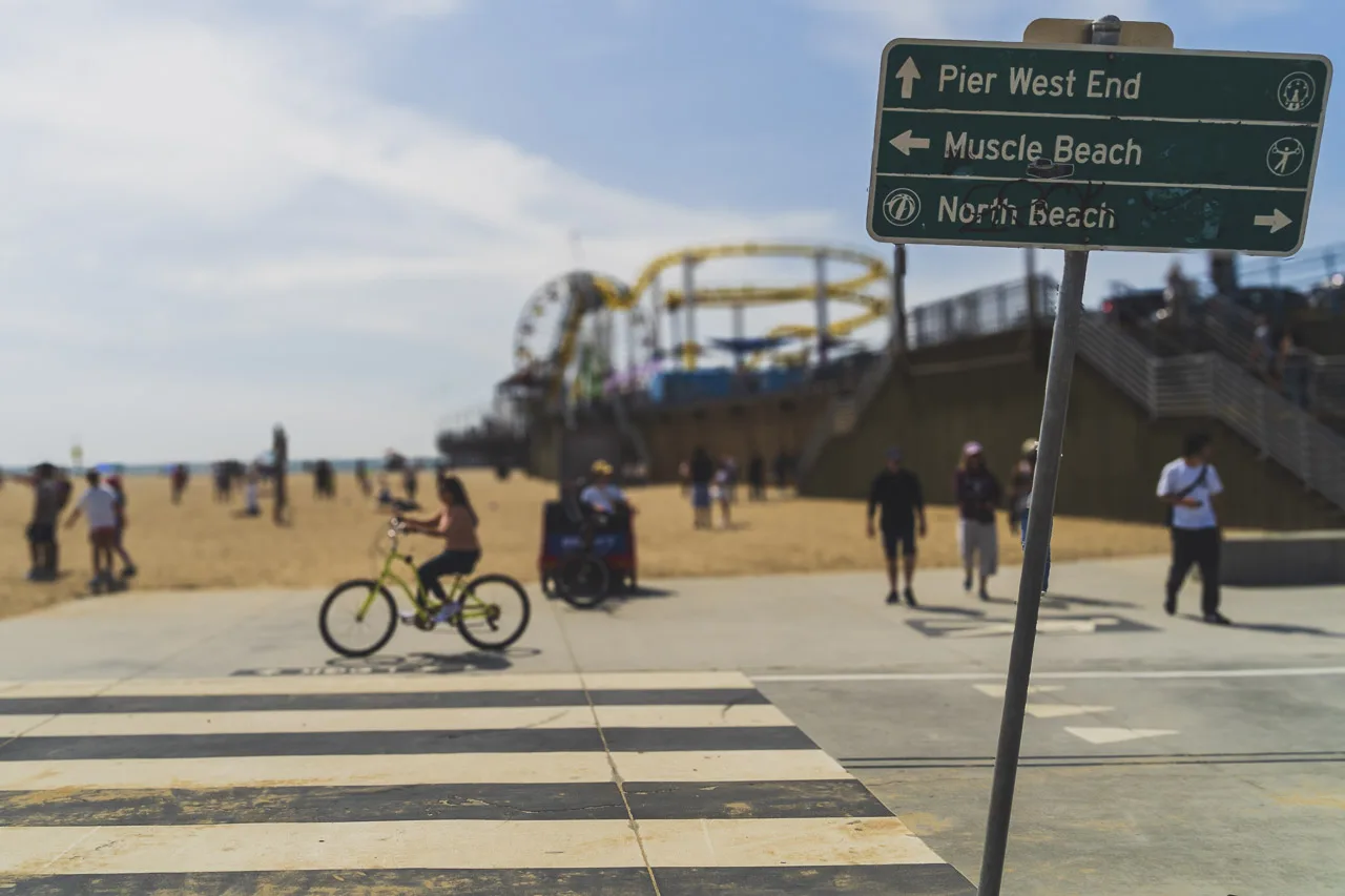 A sign says "Pier West End" pointing forward.
Muscle Beach pointing left.
North Beach pointing right.
People cycling and walking on the beach in the background as well as Santa Monica Pier.