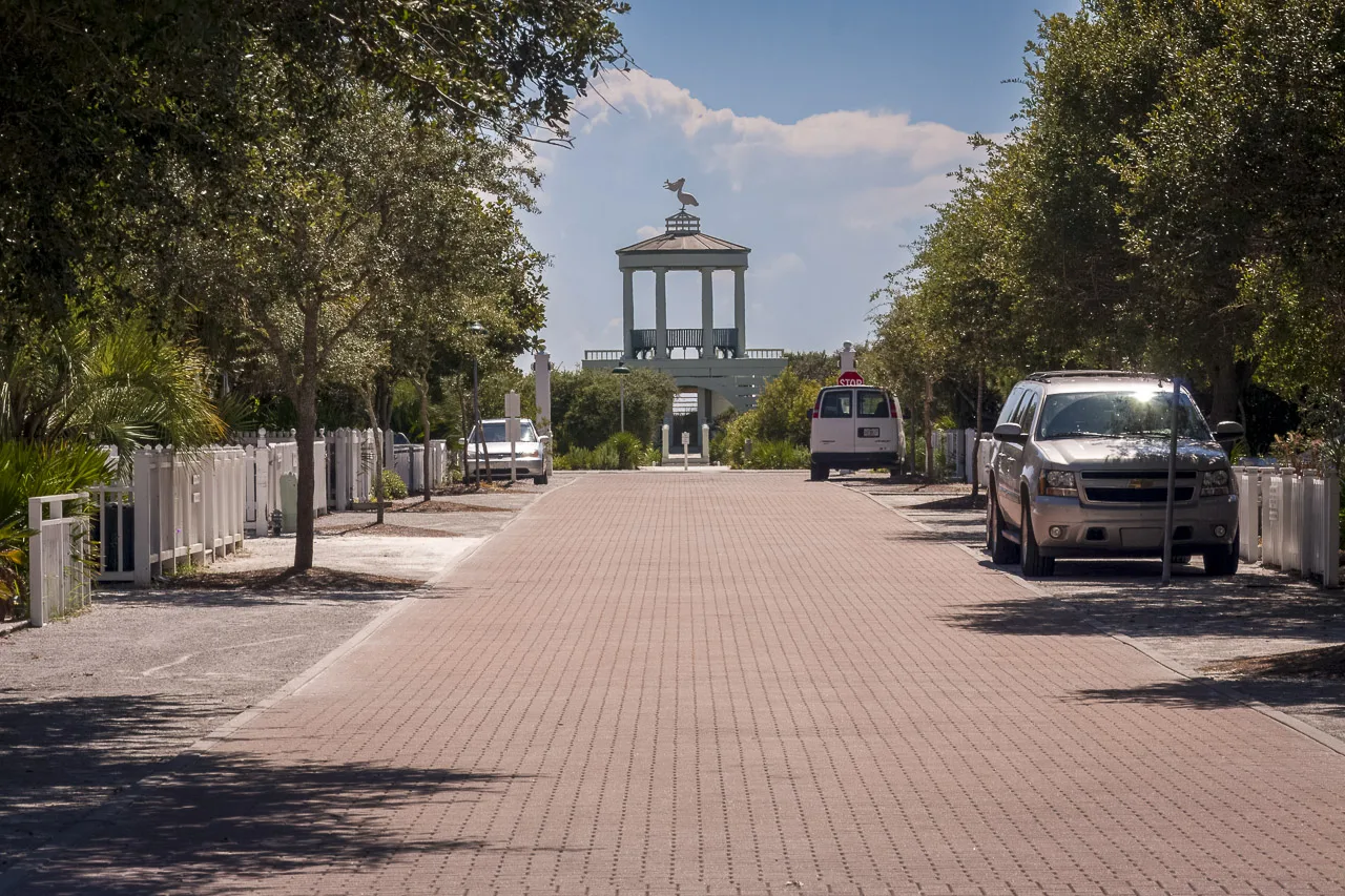 An empty road in the town of Seaside that looks very like a road in the movie The Truman Show.
The road is made of red bricks, white picket fences line the sides of the road under trees, and a few parked cars line the road.
There's an ornate building at the end of the road.