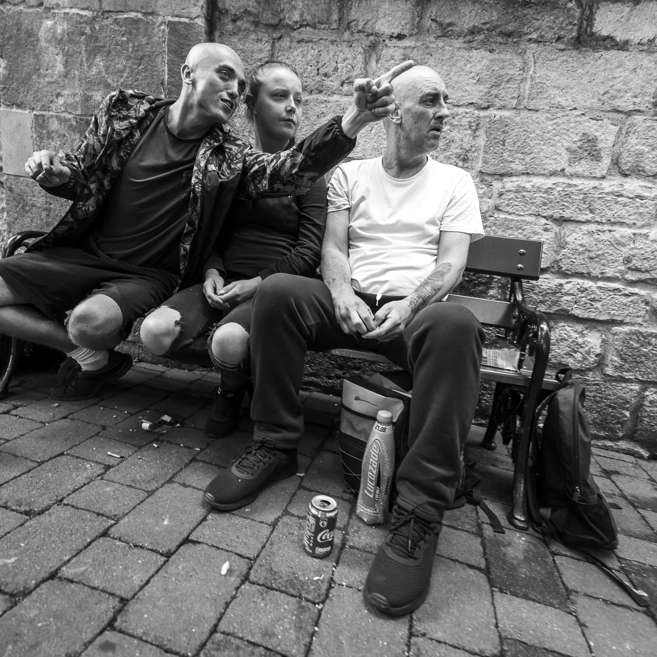 Three people sit on a bench. The man on the left points to the right and the woman sitting next to him and the other man look over.
They have a couple of bags and a few drinks with them.