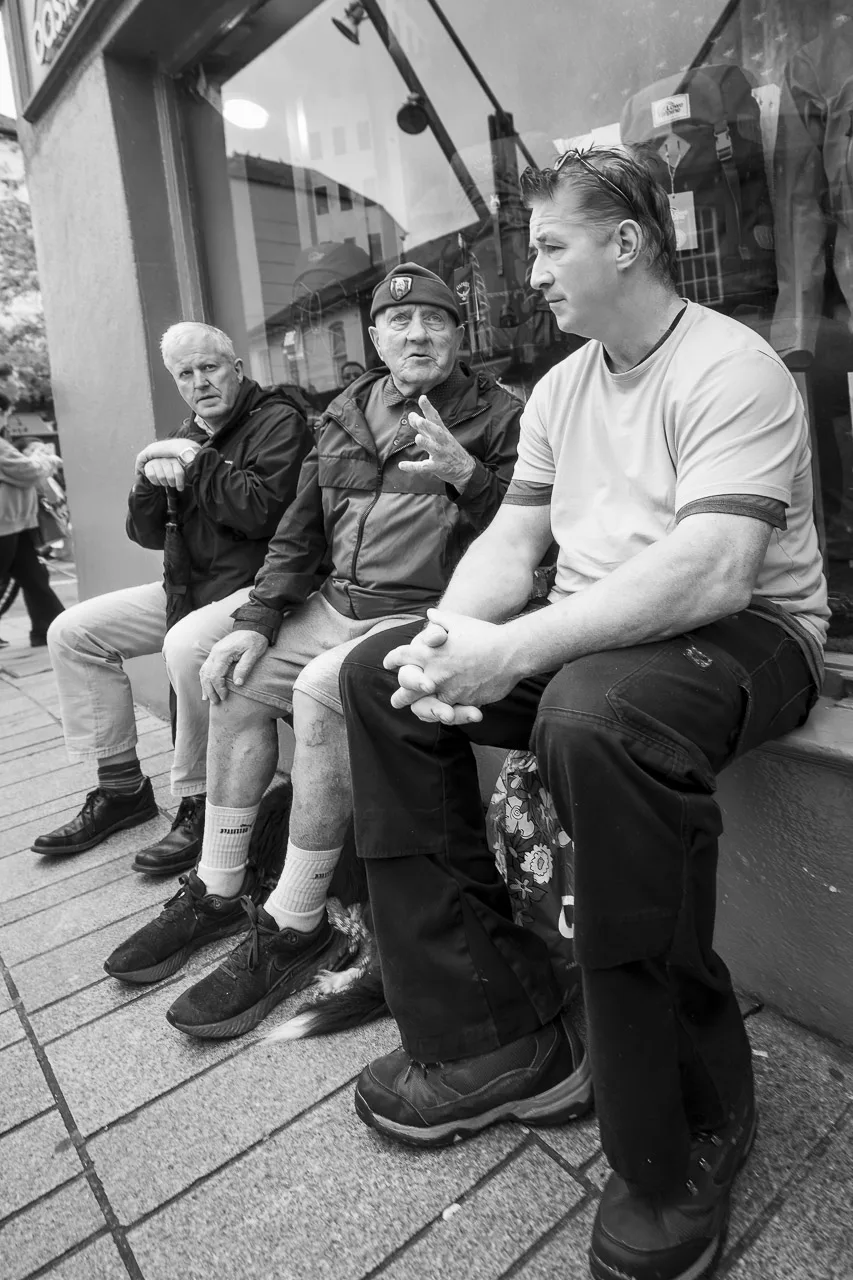 3 men sitting on a wall. From left to right, one man is looking towards the camera, holding an umbrella. The second is looking this way too but is busy talking and gesturing. A dog sits underneath this man's legs and the last man listens patiently.