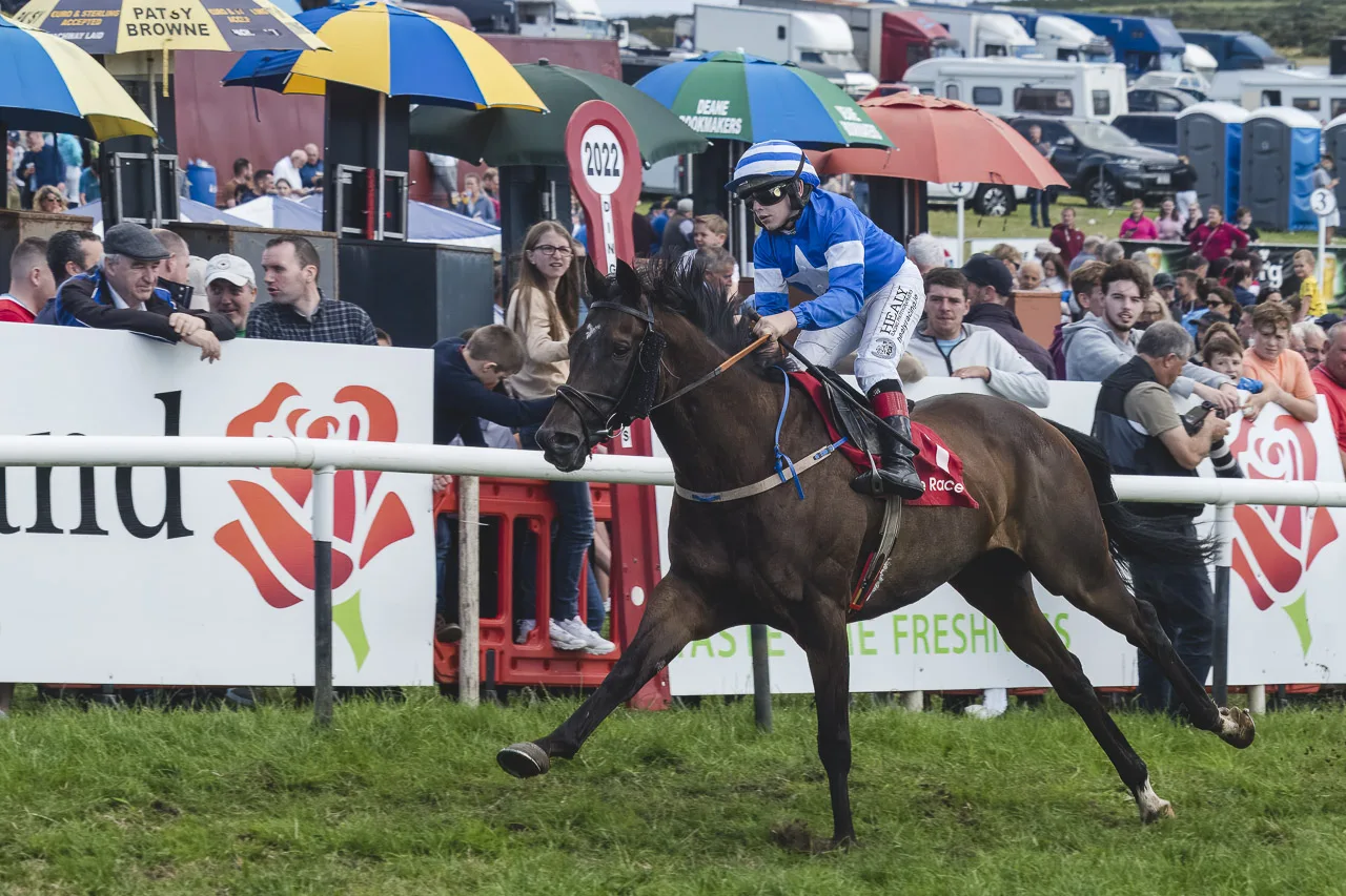 A young rider on a brown horse races past the finish line after doing a lap of the track on grass. People watching from behind the barrier and umbrellas offering shade to bookies behind.