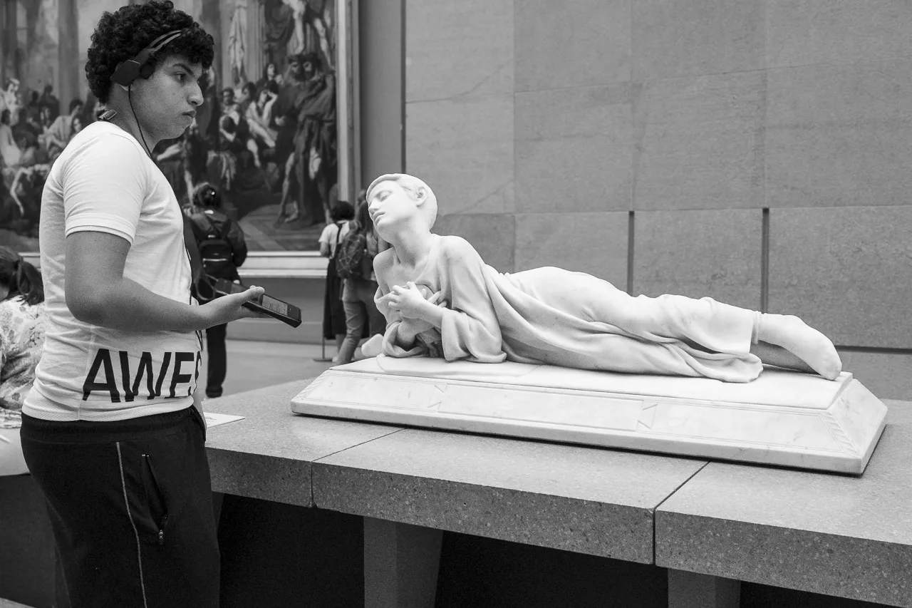 A young man listening to the audio tour of the Musée d’Orsay in Paris stands next to the statue of Tarcisius.

The young man's tshirt says "AWE" and what is probably "S", leading me to believe it says "AWESOME".