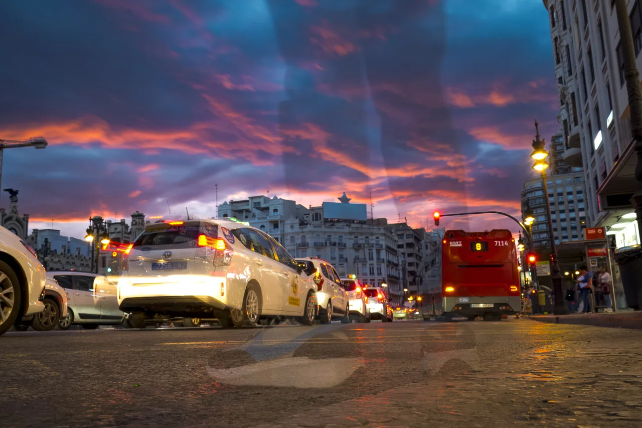 In a long exposure photo of a street in Valencia, someone stood in front of my camera so their legs are seen as transparent shadows in the photo. Busy street with taxis and a bus under a cloudy sky lit by the setting sun.