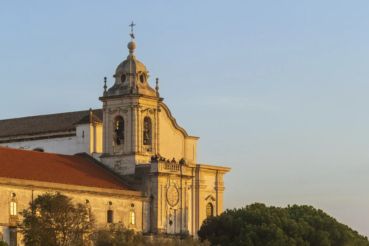 The setting sun casts a yellow glow on a church with a clear blue sky behind it. People are standing on a balcony watching the sunset.