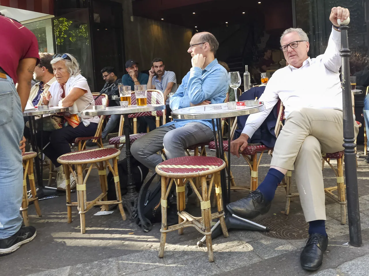 People sitting at tables outside a café in Paris, France.
Three men sit at tables in the background, while two men sit at tables nearer, and a couple are at another table, being waited on by a waiter.