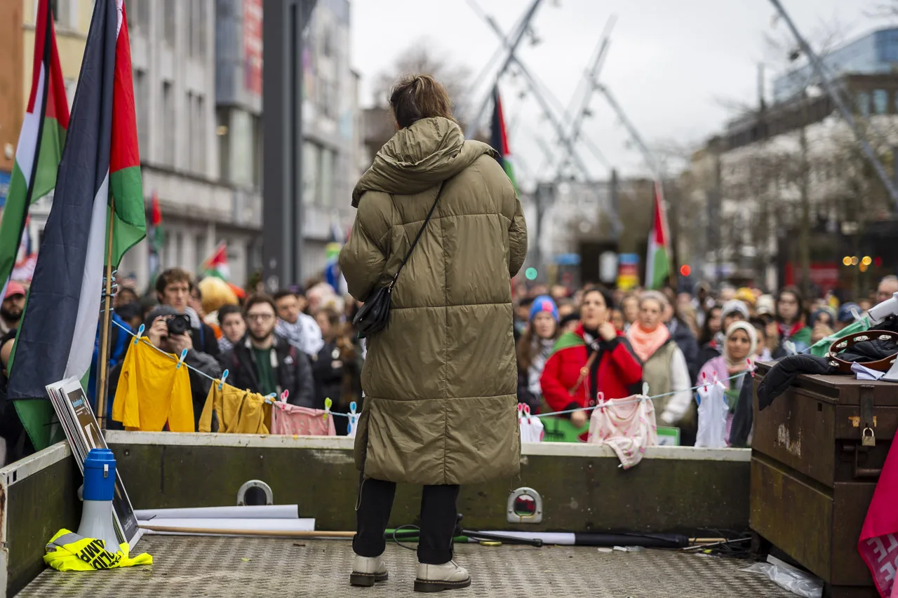 A large crowd watches a woman talking. She is pictured from behind and is wearing a coat on a cold day. The crowd is out of focus, listening to every word.