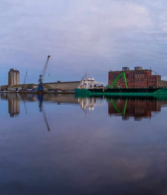 A wide view of Kennedy Quay, going on to Albert Quay in Cork. Buildings are reflected in the River Lee and a green ship is seen docked on the quay. (1/3)