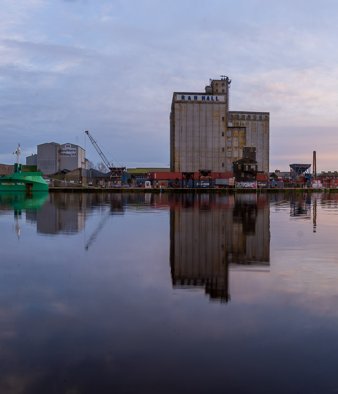 A wide view of Kennedy Quay, going on to Albert Quay in Cork. Buildings are reflected in the River Lee and a green ship is seen docked on the quay. (2/3)