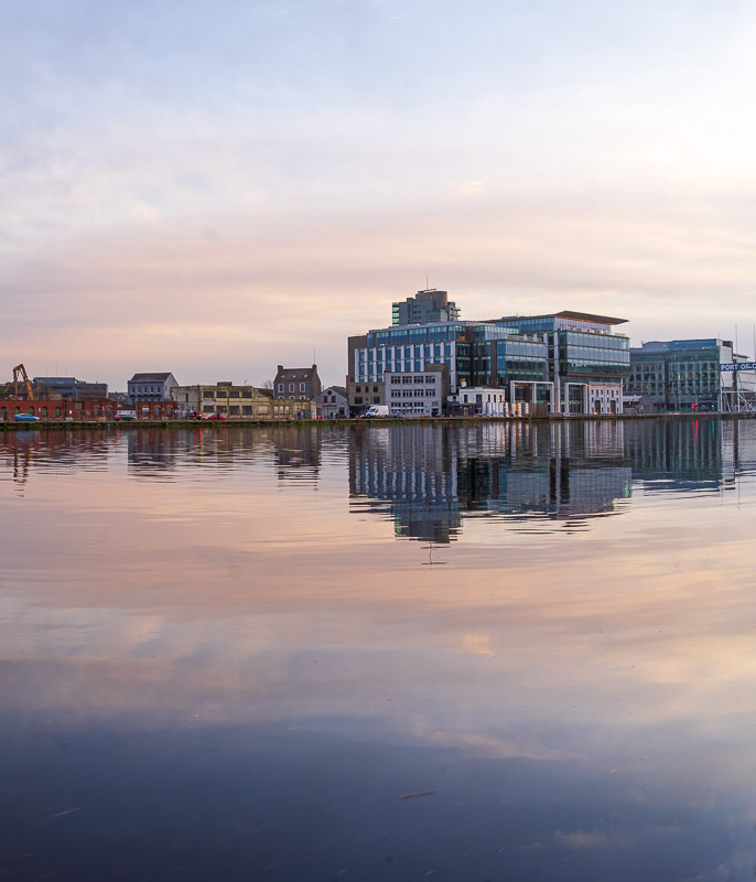 A wide view of Kennedy Quay, going on to Albert Quay in Cork. Buildings are reflected in the River Lee and a green ship is seen docked on the quay. (3/3)