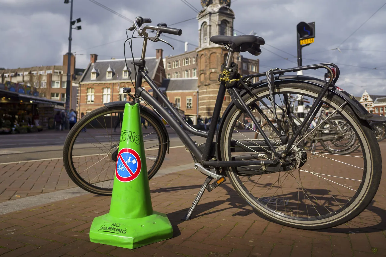 A bike parked at the side of the road. It's stand down and no rider nearby. A green code sits in front of it saying "NO PARKING" and a sign that indicates no bike or scooter parking.