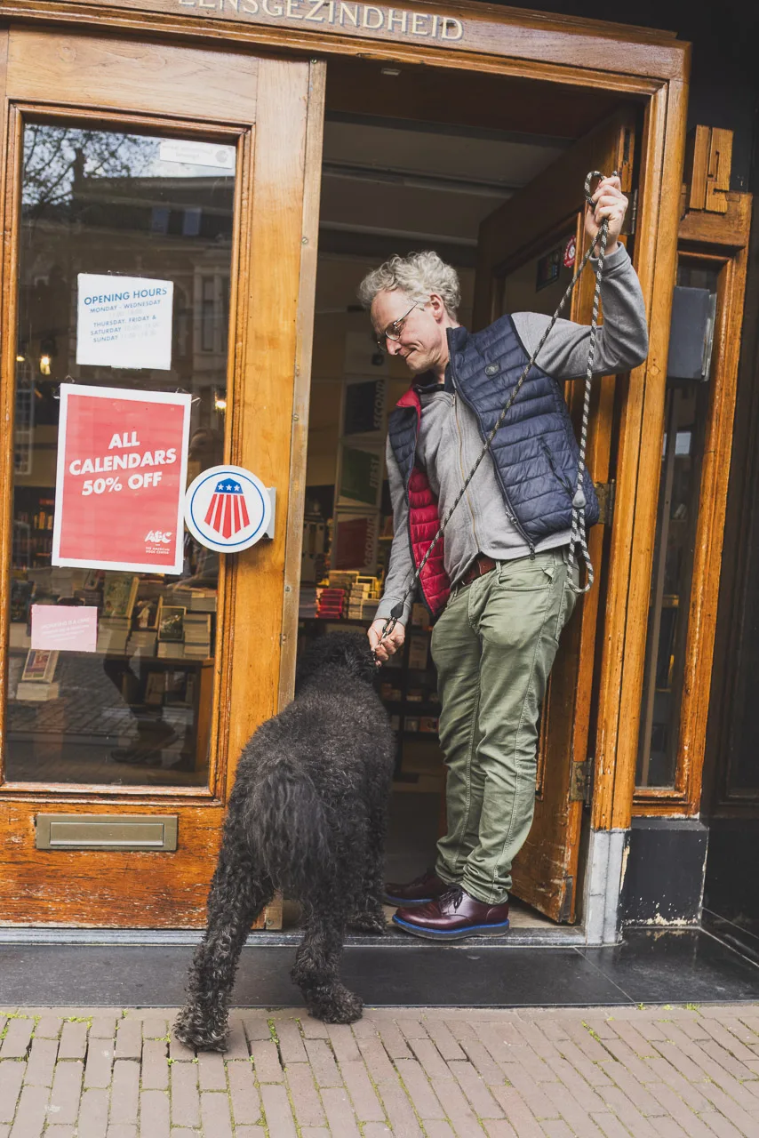 A man stands in the doorway of a bookshop next to his large dog. He holds the lead but looks like he is about to remove the lead as he goes in!