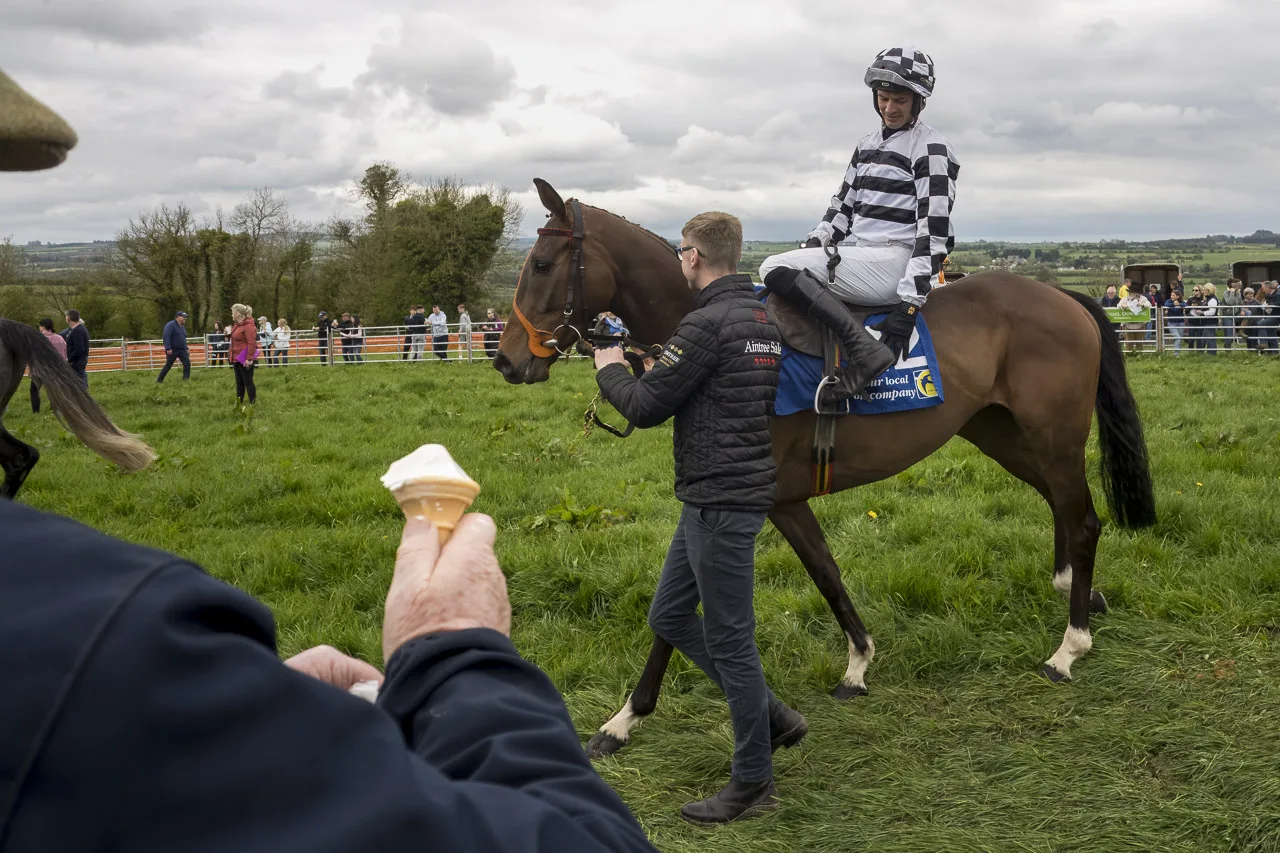 A jockey sits on his horse, putting his foot in the stirrup as another man guides the horse. The arm of a spectator is seen in the foreground, holding an ice cream.