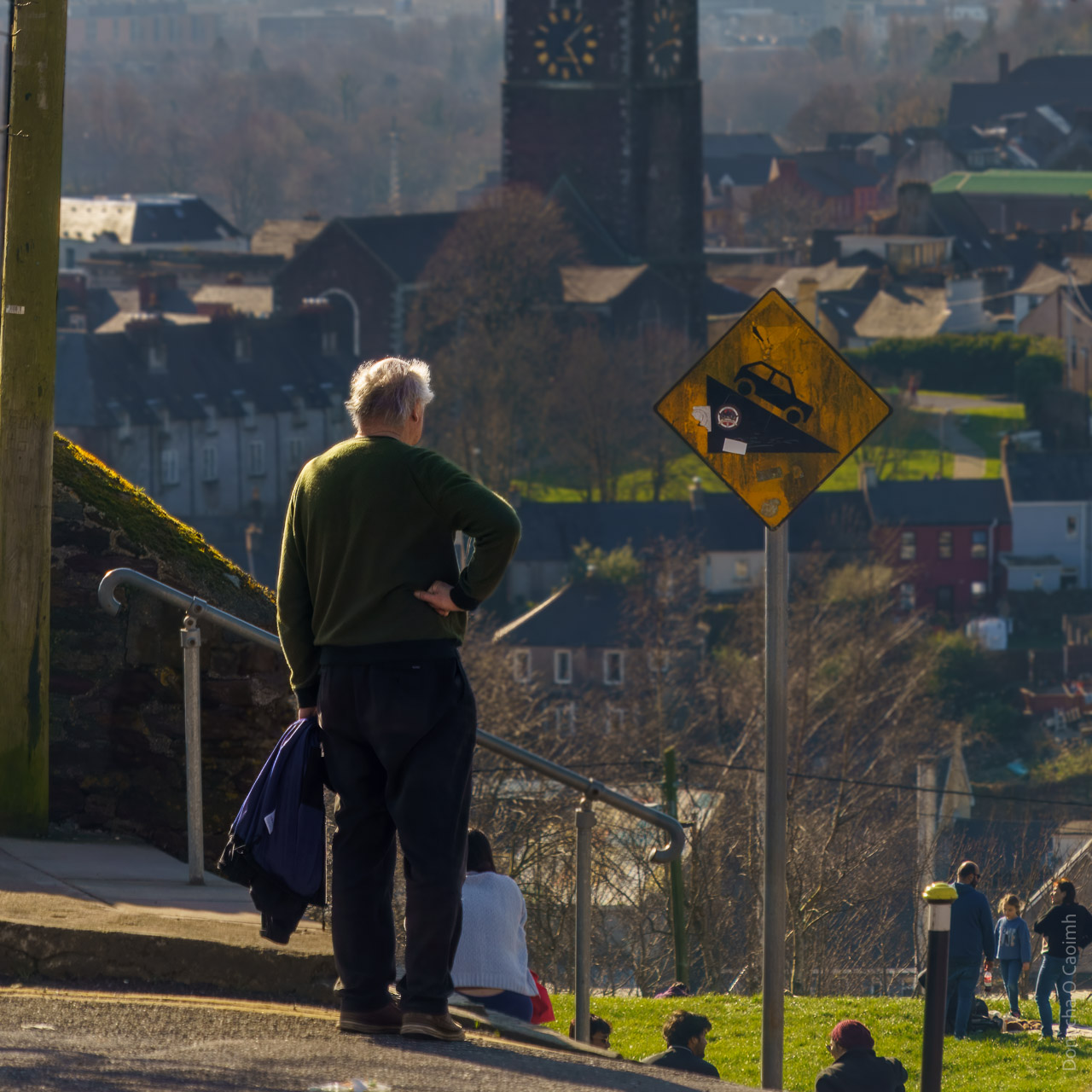 A man looks out on the North Side of Cork, in front of a steep road. A sign showing a car on a steep incline stands in front of him.
