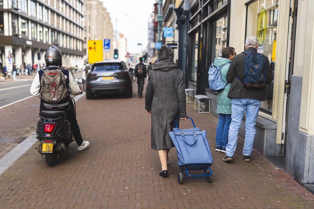 A moped rider, a shopper with a bag and two people looking in a shop window all in their own worlds together on the street. Up ahead a car is parked illegally on the pavement.