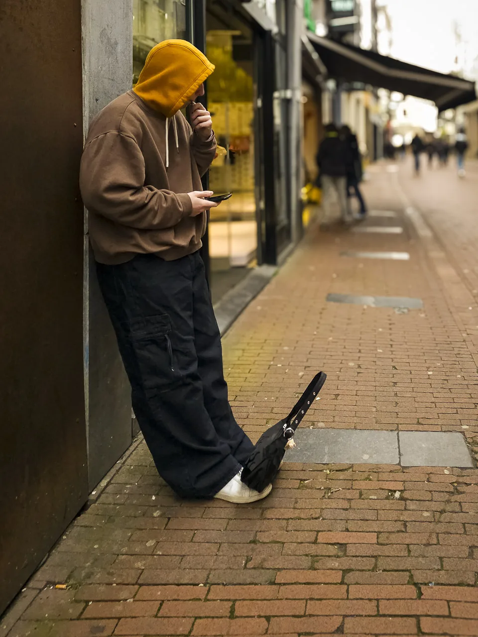 A person leans against a street wall wearing a very baggy hoody and trousers. They are vaping with one hand and holding a phone in the other. A handbag sits on their feet.