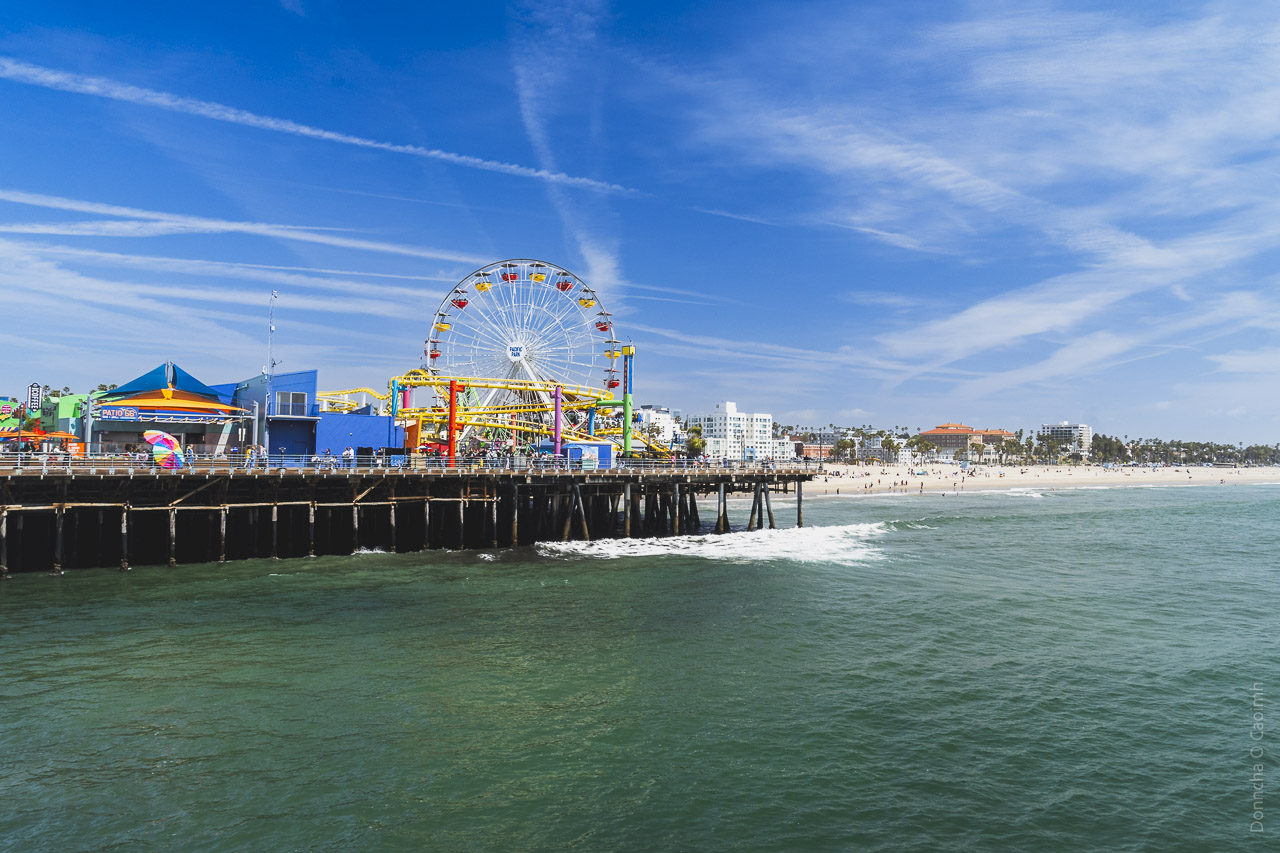 Looking back on the pier in Santa Monica and the funfair and rides, including a ferris wheel. Further back is the sandy beach and coastline. A blue sky with faint cloudy lines.