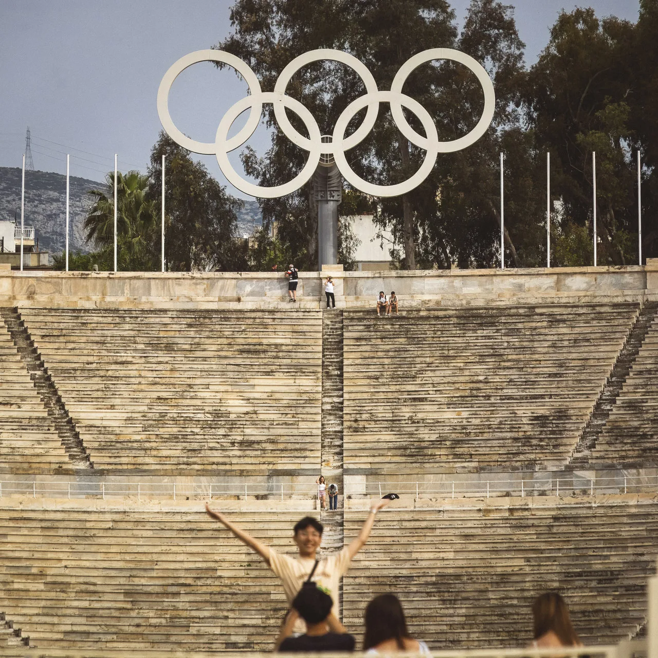The Olympic Rings are clearly visible over the Panathenaic Stadium in Athens. A few people are sitting of walking around the steps of the stadium and someone is in the near foreground smiling for the camera as his friend takes a photo of them.