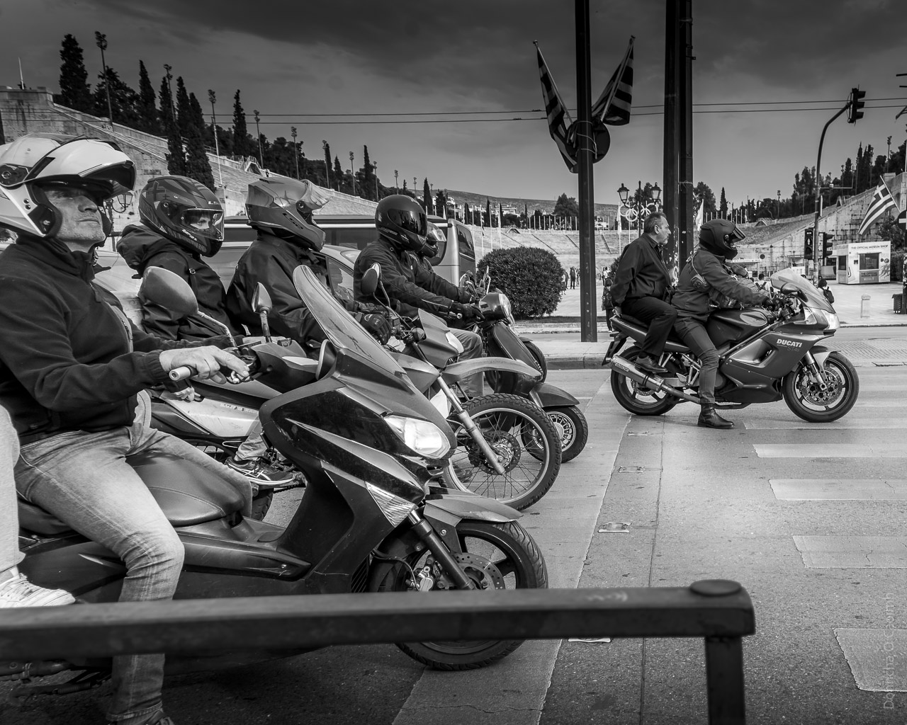 Motorbikes stopped at traffic lights in front of the Panathenaic Stadium in Athens. A rider looks off in the distance with the front of his helmet open. He has a passenger as a leg and foot is just visible behind him.
Four other riders are spread out across the road and the furthest away bike has a bare headed passenger.