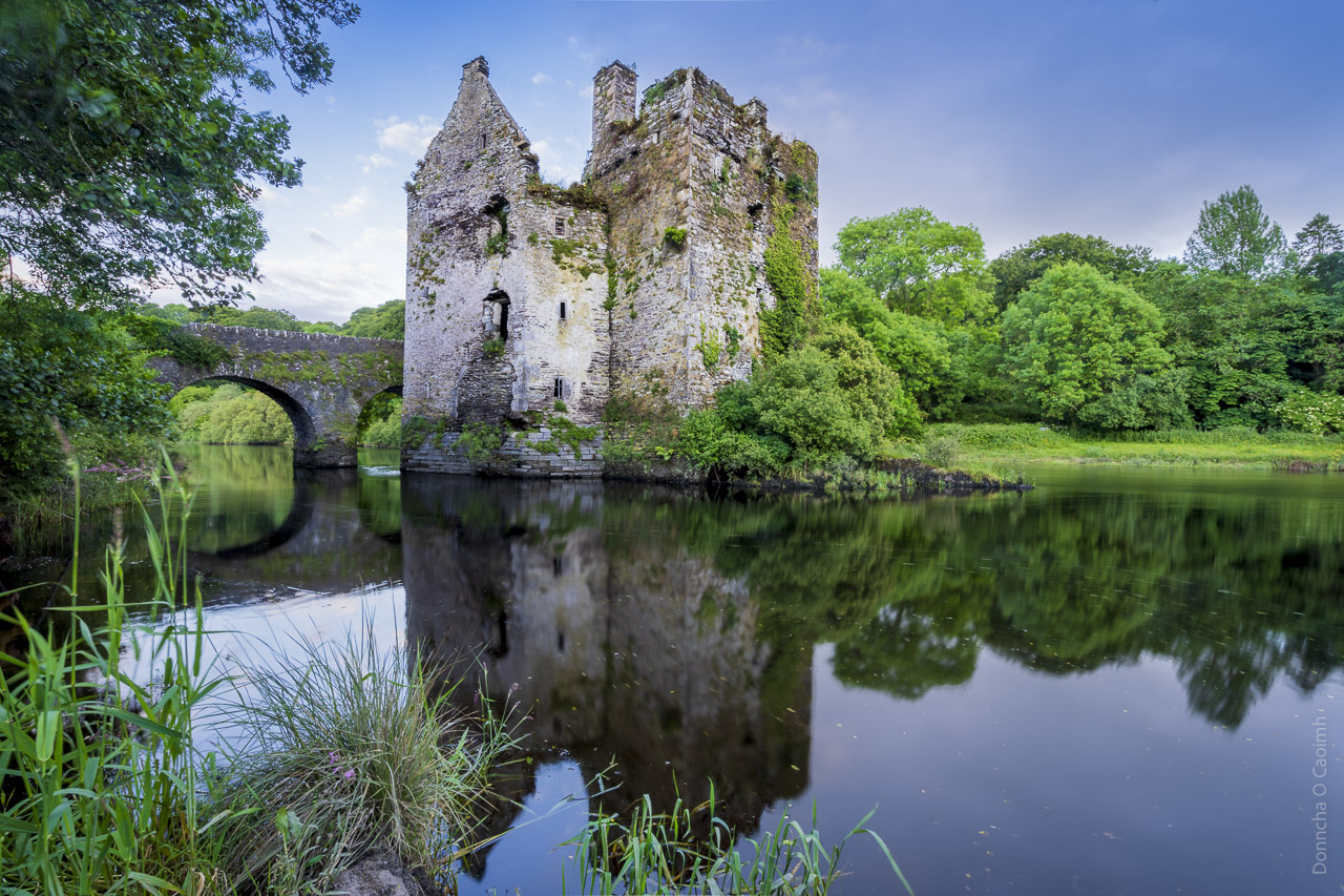 A ruined castle standing in a river next to a bridge. The castle and bridge are reflected in the calm waters of the river while trees line both banks.