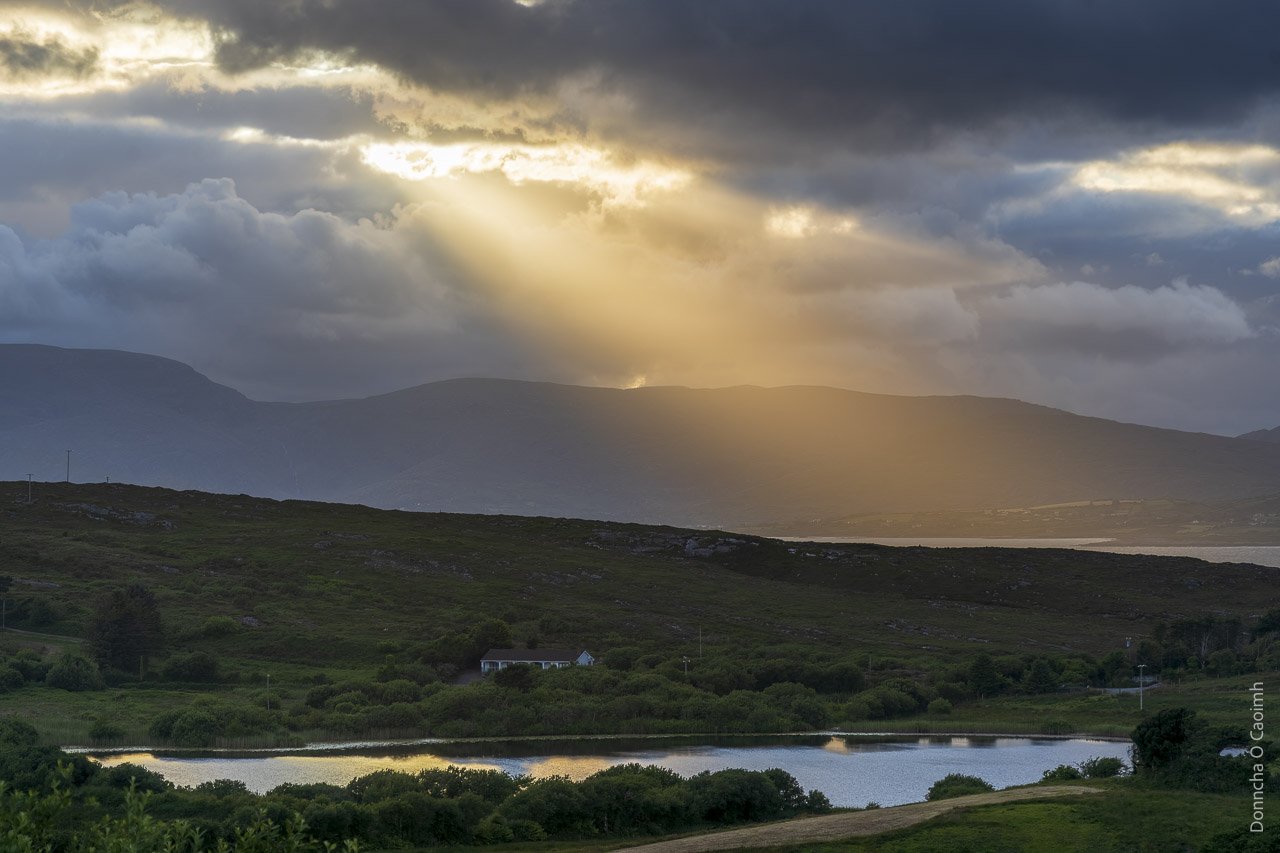 Light pours from a gap in the clouds near sunset over Sheep's Head. There's a small lake in the foreground, reflecting the light, and a bungalow sits near the lake.