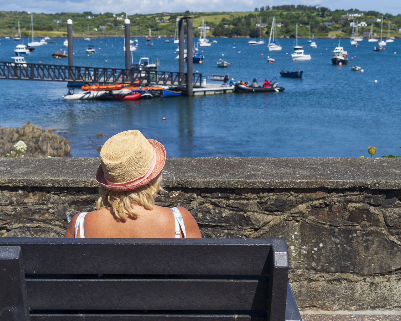 A woman sits on a bench, seen from behind. She's wearing a hat and watching the harbour, where there are many boats and a few people manoeuvring one boat on to a dock.