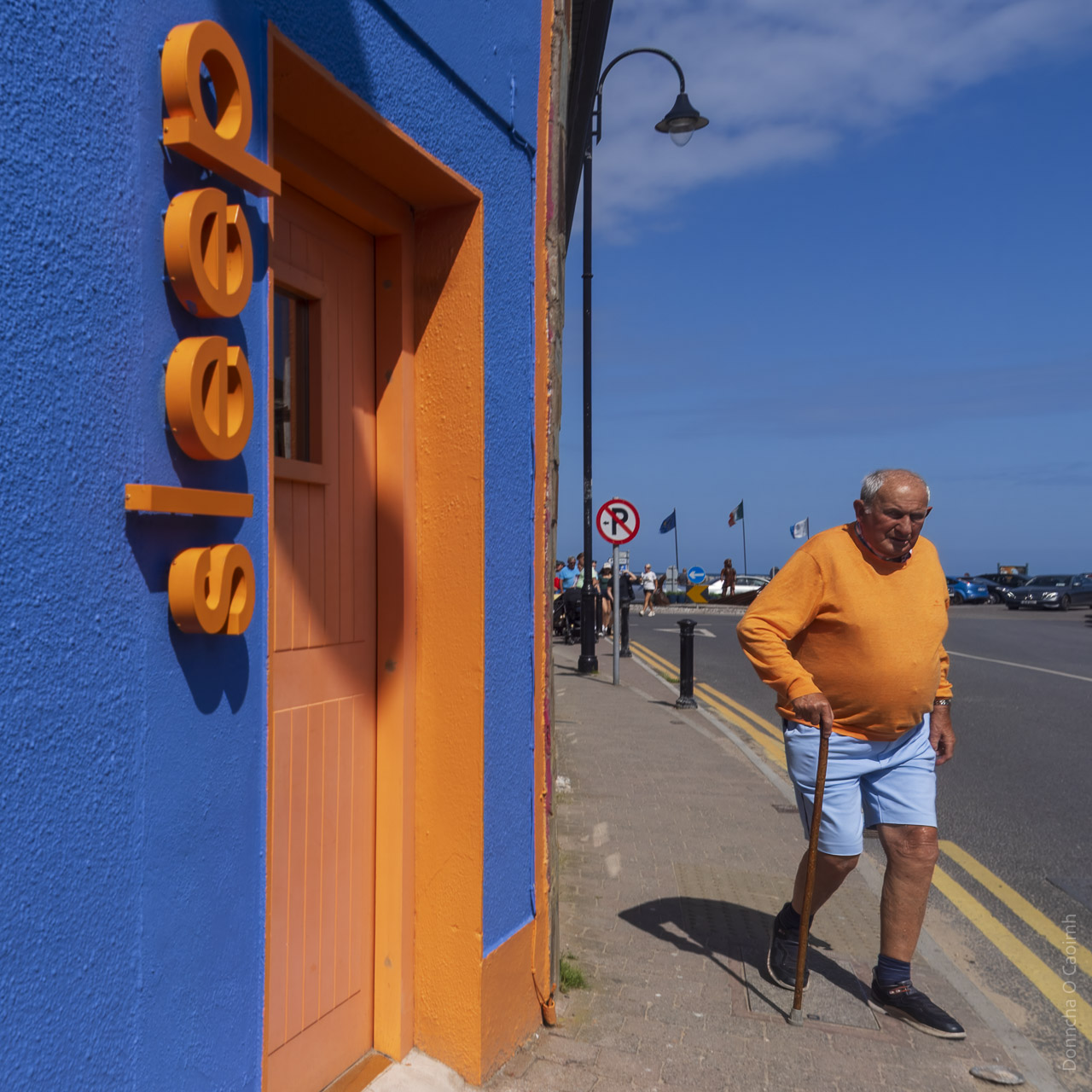 A man dressed in a bright orange sweater and light blue shorts walks, with the help of a walking stick, past a hostel that is painted bright orange and dark, deep, blue. Letters forming the word, "sleep" are on the wall, next to a doorway. Both are in the same bright orange shade.