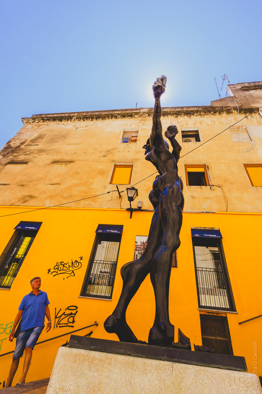 One of Salvidor Dali's statues in Figueres as seen from below with a man in the background looking up. A yellow building stands on the other side of the narrow street.