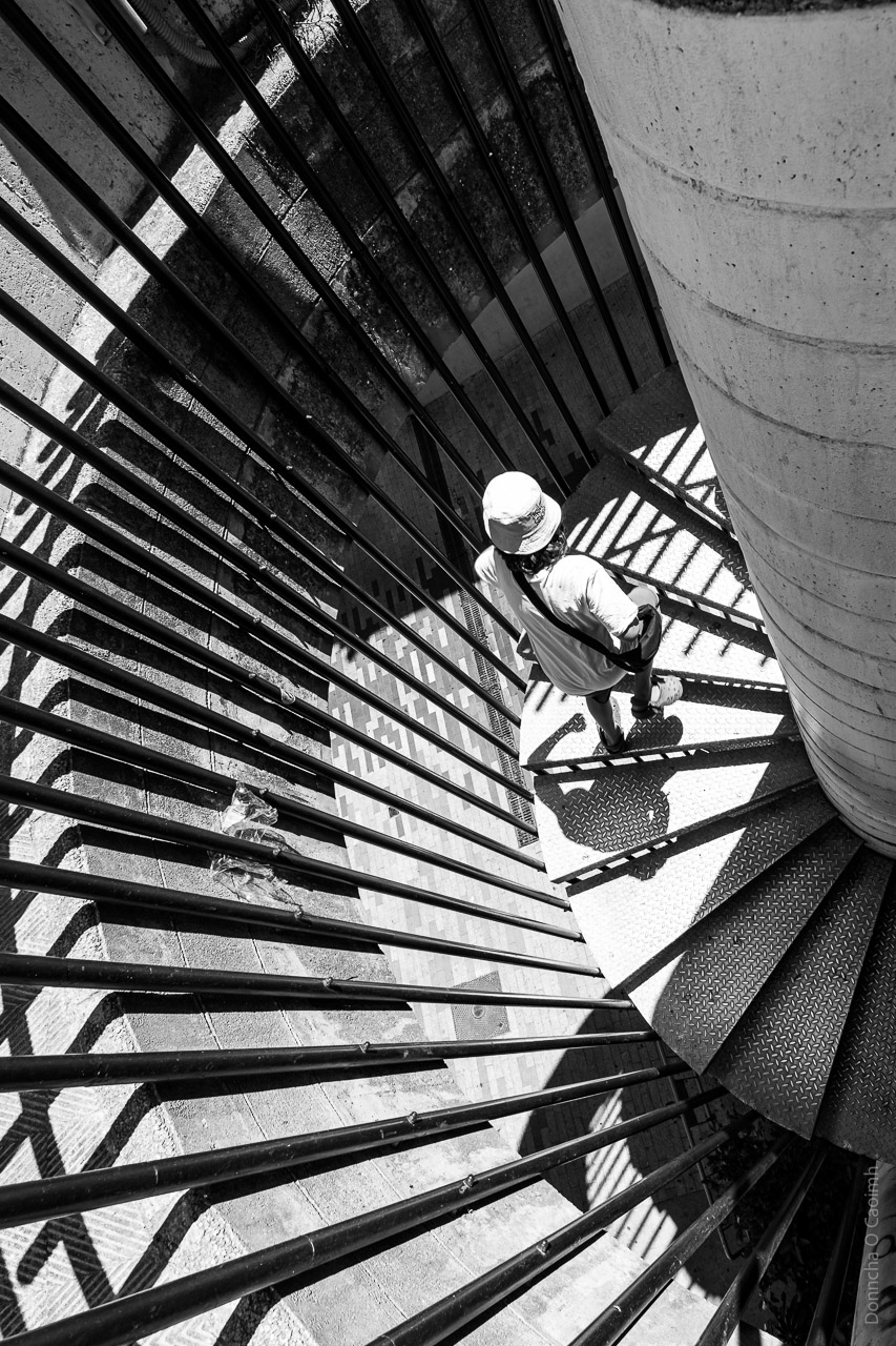 A person wearing a hat on their head (and the usual clothes) climbs the circular stairs at a multi-storey car park.