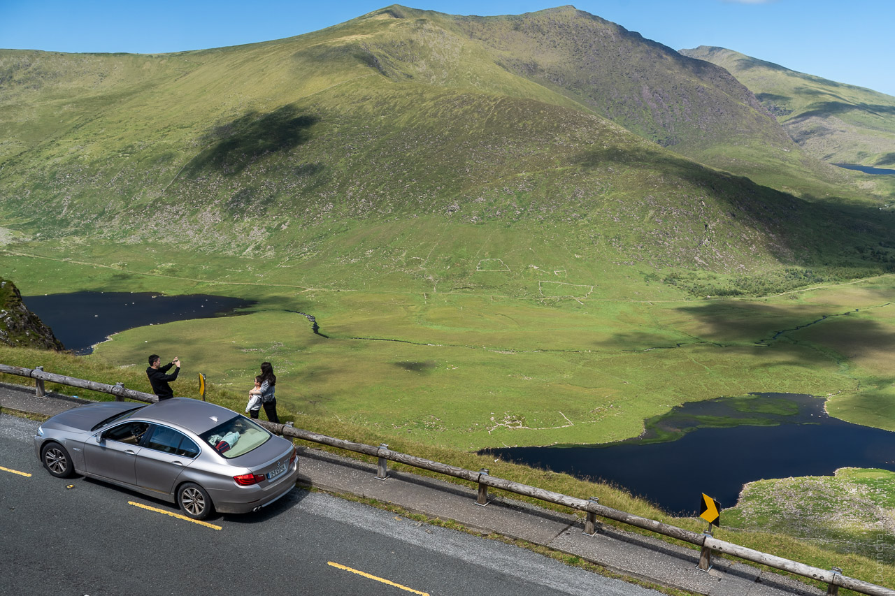 A family taking photos at the side of the road. Their silver BMW car is pulled in next to them while they take photos with the backdrop of the magnificent Conor Pass behind them.