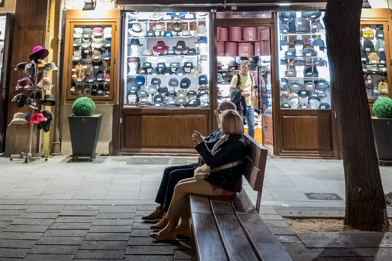 A couple sat on a bench while a man exits the door of a hat shop. There are LOTS of hats in the windows of the shop.