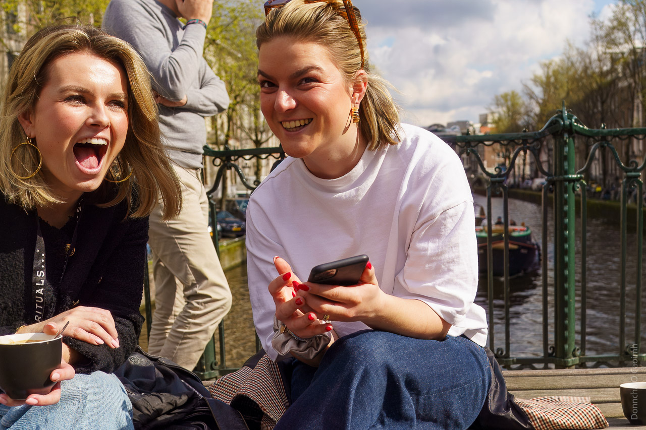 Two women seated on a bench on a bridge in Amsterdam laughing and chatting. One holds a mug, the other a phone. A man walks behind, and a barge is visible on the canal in the background.