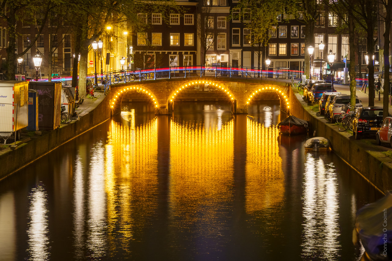 Lights shine from a bridge over a canal in Amsterdam. The light trails of a passing vehicle add to the light on the bridge.