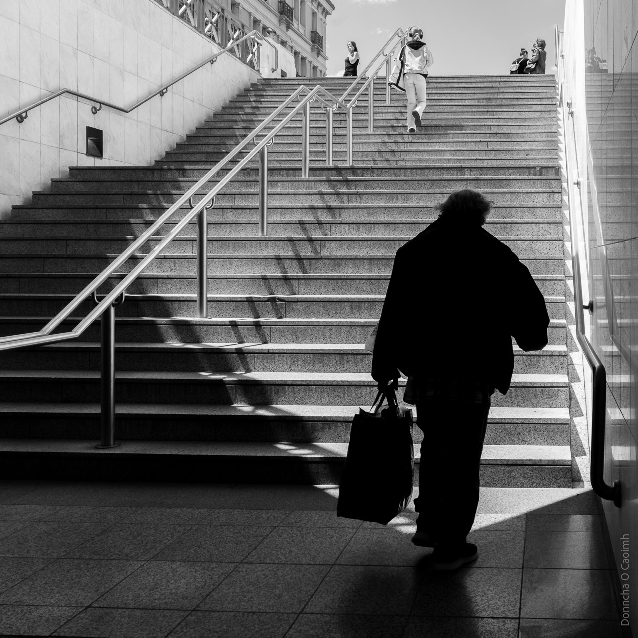 The silhouette of a man as he is about to walk out of the shadows in a metro station in Athens, Greece.
Stepps around in front of him with a few people visible further up.