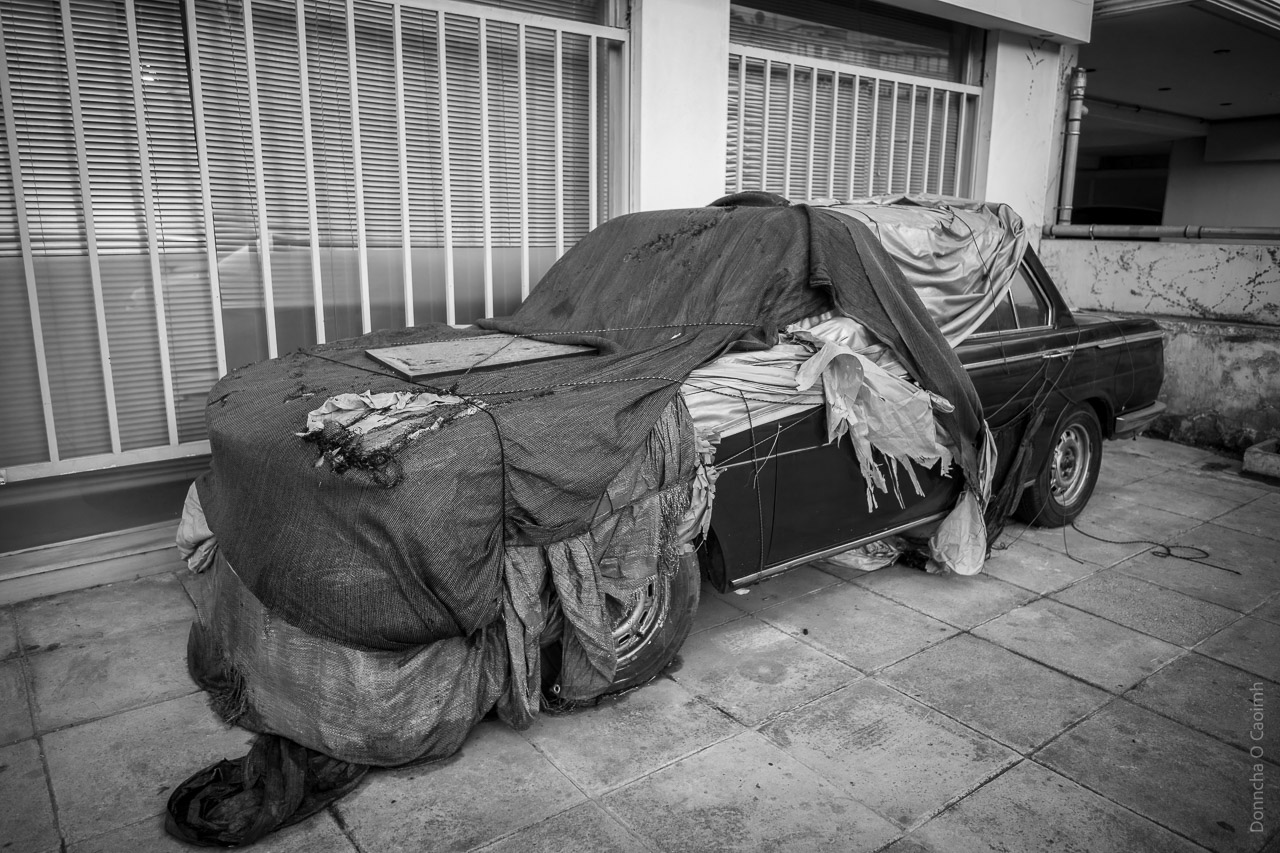 a car half covered in cloth and plastic parked on a pavement next to windows that are barred with railings. It is half-covered.