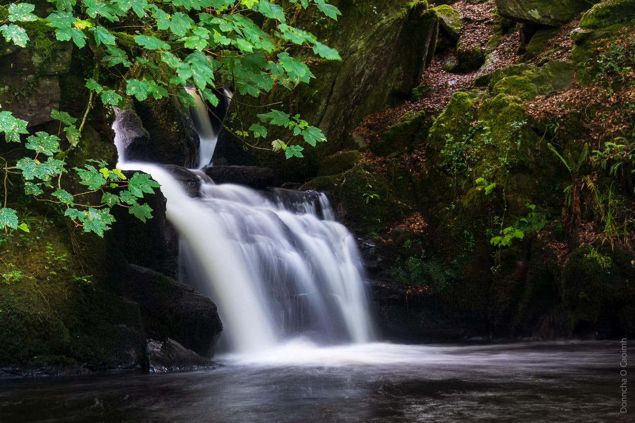 The waters of the waterfall in Mullinhassig flow in a long exposure blur into water at the bottom. Surrounded by mossy rocks and leaves.