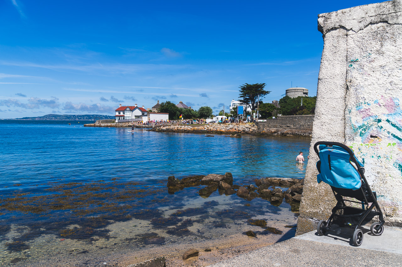 A buggy sits on the concrete next to the sea in Dun Laoghaire. In the distance is Sandycove which is thronged, and a man is visible in the near distance in the water.