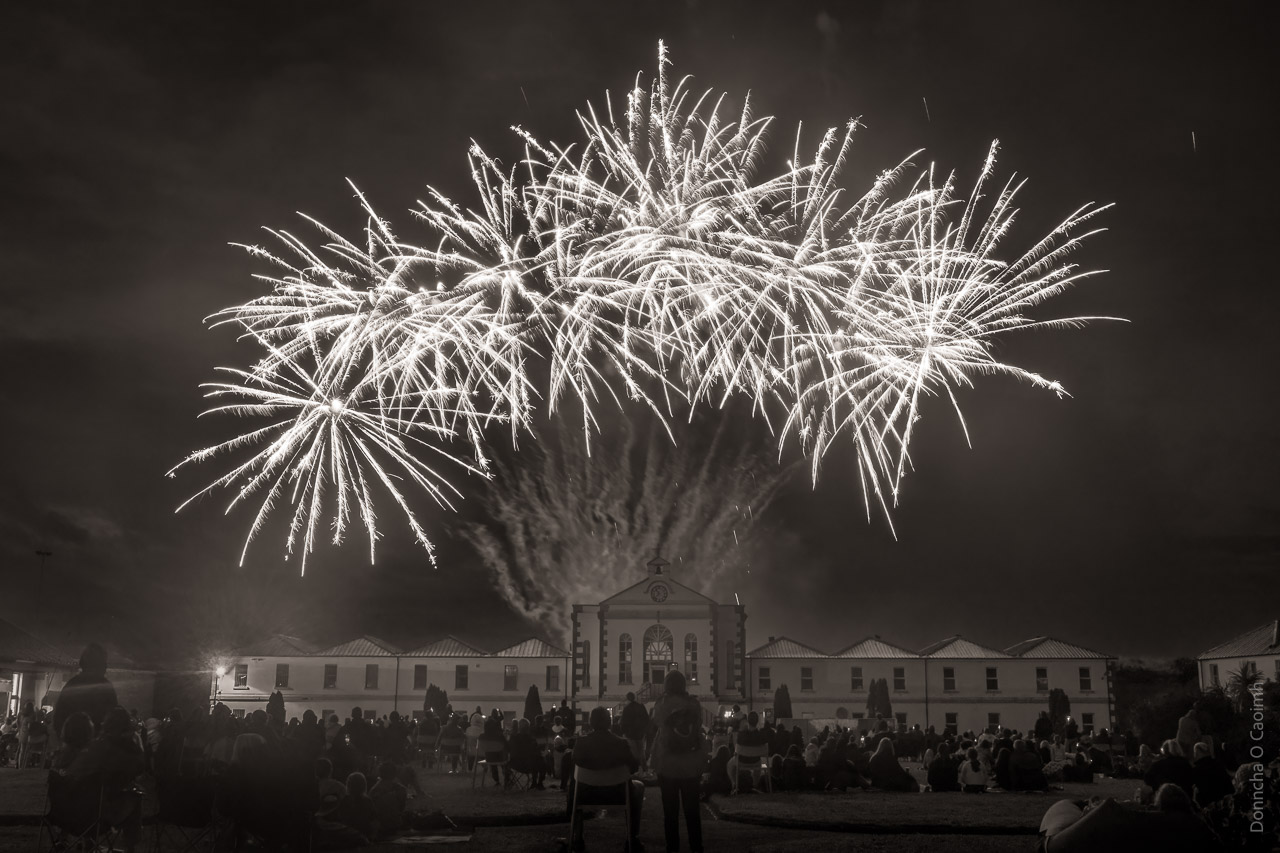 A black and white image of fireworks over Spike Island. Buildings are seen in the background and people are sitting on the ground watching.