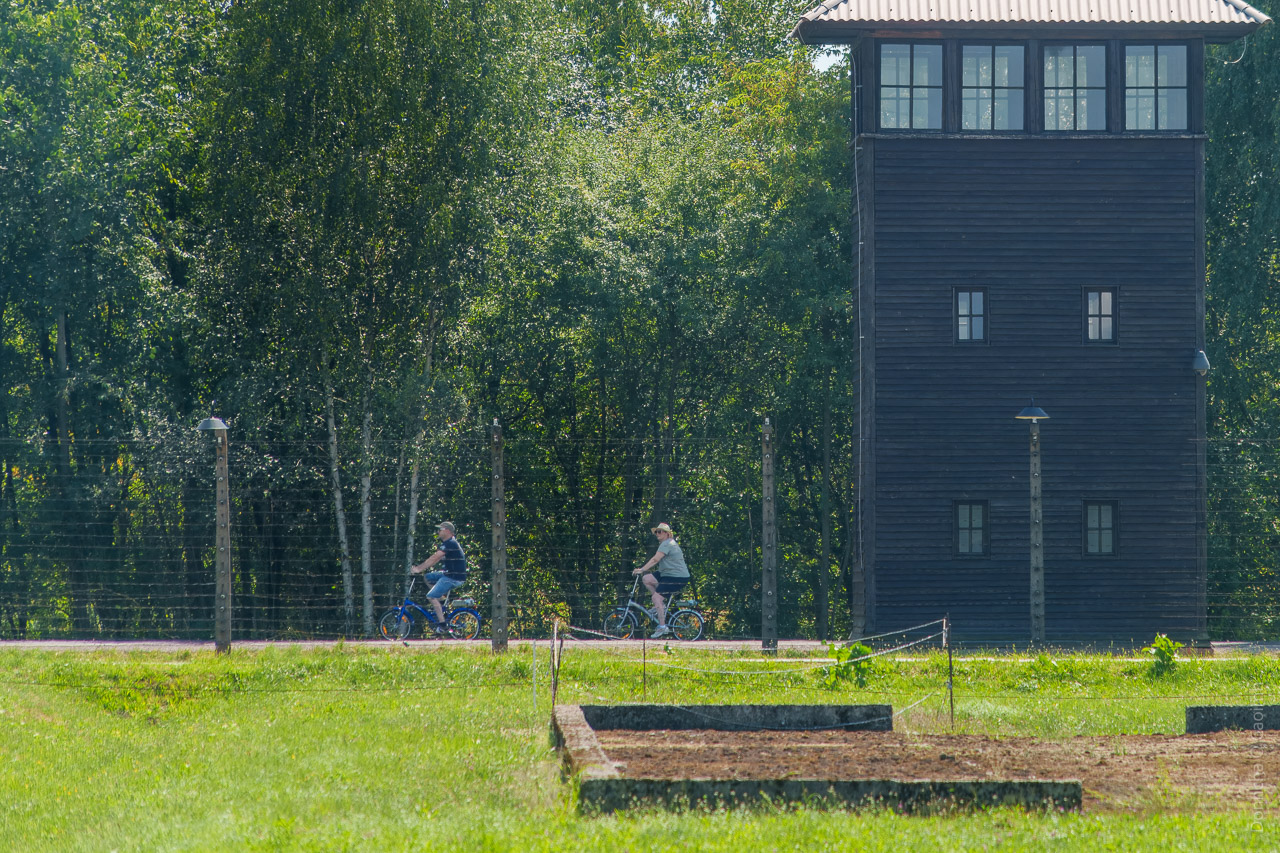 Two people cycling on the other side of a barbed wire fence with a watch tower to the side.
A derelict site is seen in the foreground.
