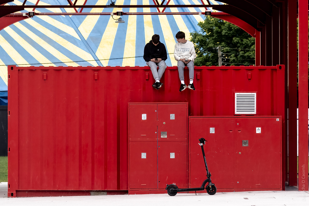 Two teenagers sit on a red container. On their scooters is visible on the ground below, and a circus tent can be seen in the background.