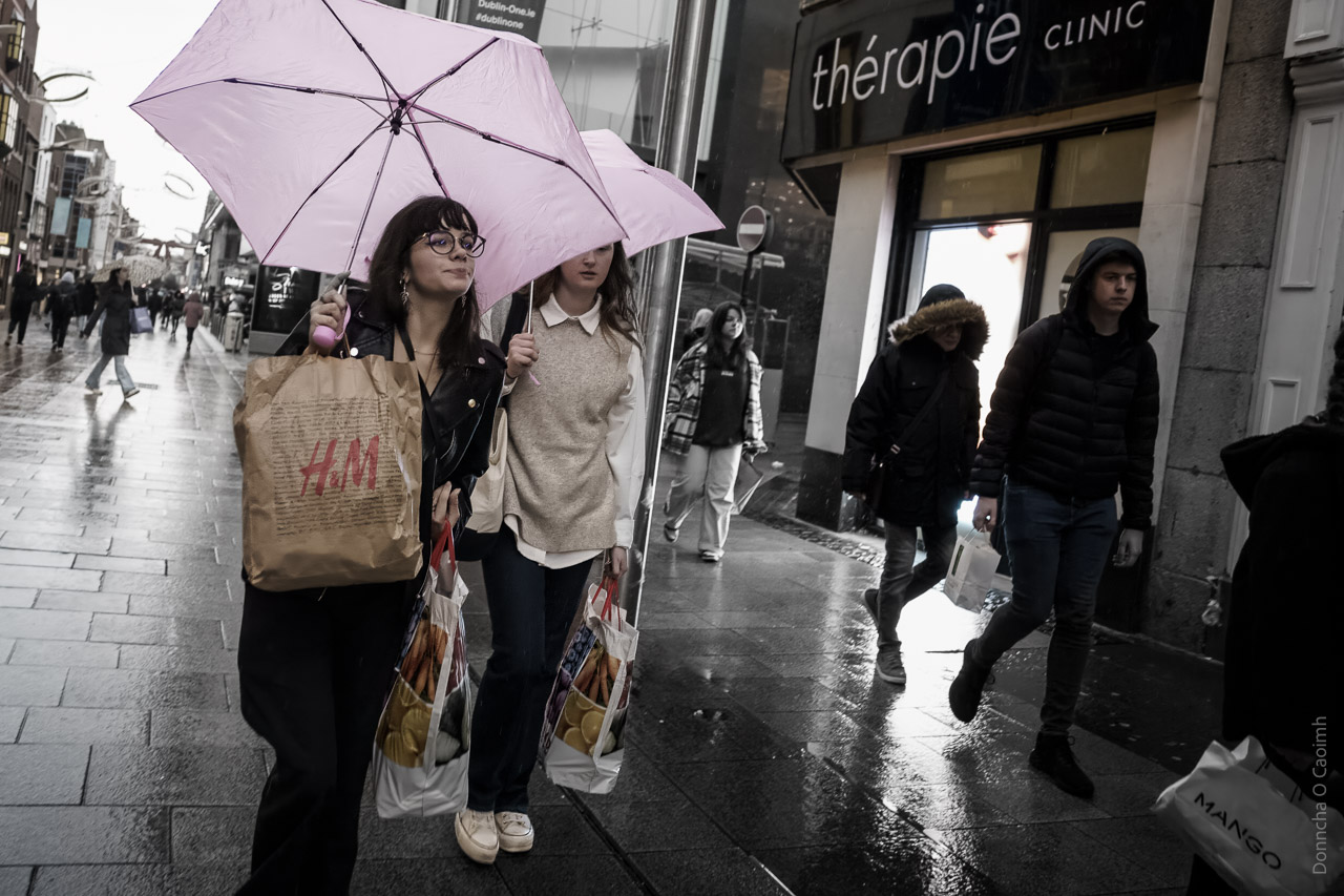 People walking on a wet street while it is raining. Two women have umbrellas and carry shopping bags.