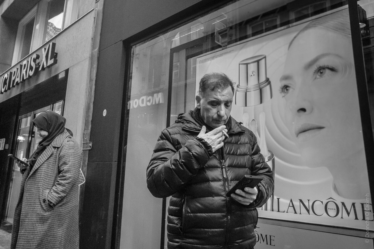 A man smokes a cigarette while looking at his phone while a woman in the background looks at her phone. A Lancôme poster in a window shows a young woman's face, flawless and forever young.