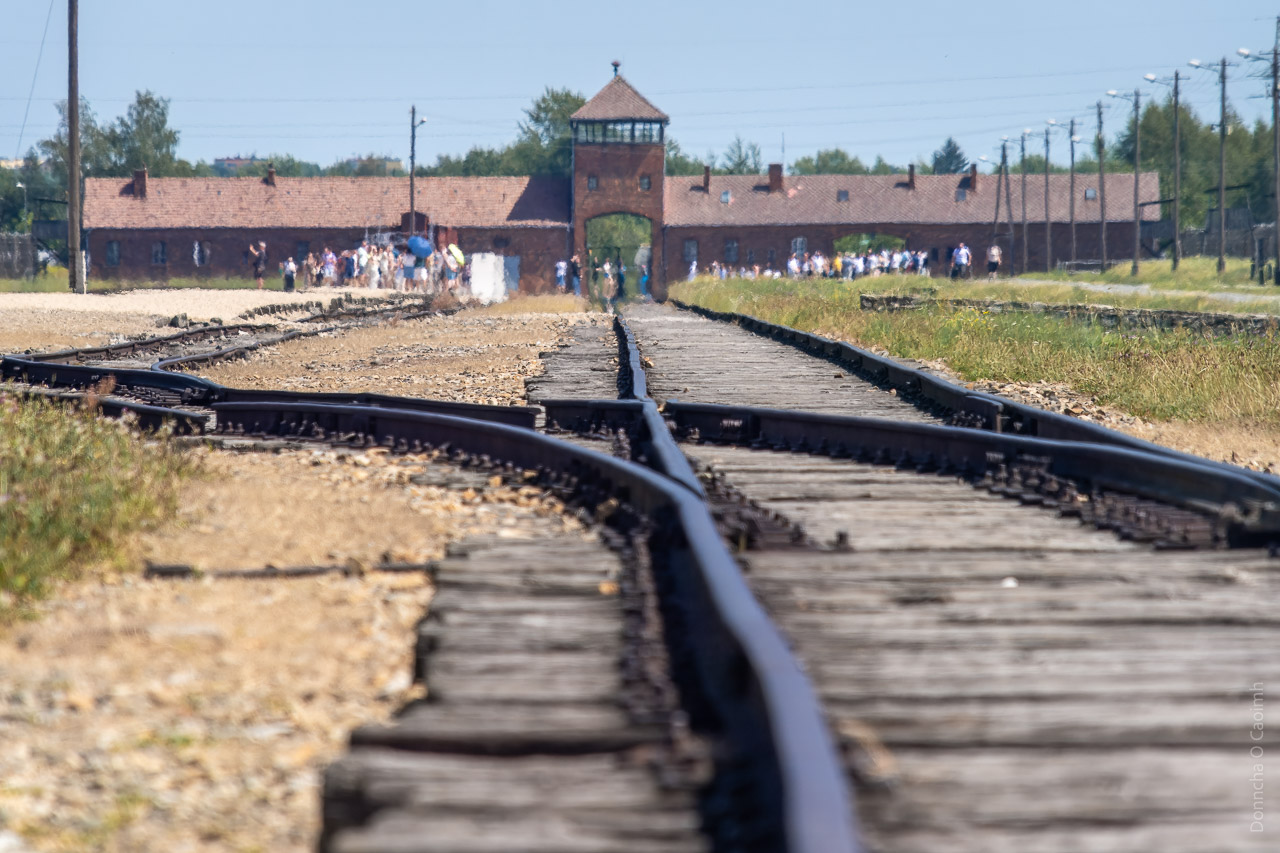 Train tracks lead off into the distance to the front gate of the former concentration camp at Birkenau. Groups of tourists can be seen congregating around the building